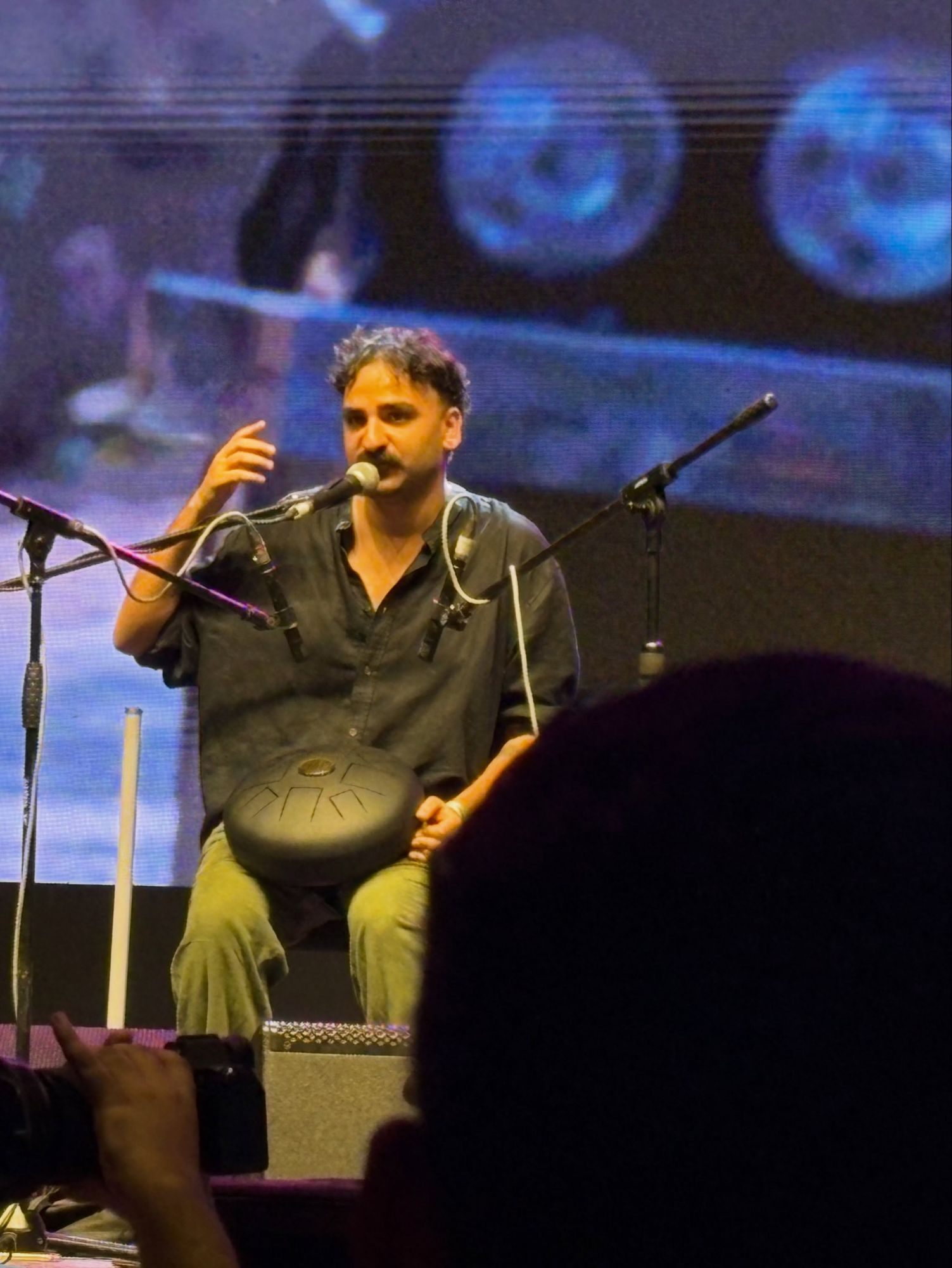 Ashim Bery seated on a large stage with a tongue drum, speaking into a microphone, a projection behind him and the audience in the foreground
