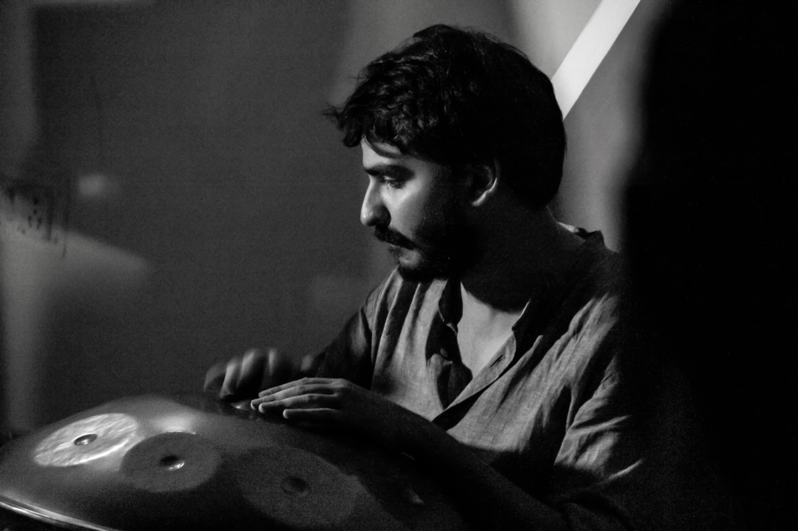 Black-and-white photo of Ashim Bery in profile, looking down at his handpan as he plays, light falling across the steel