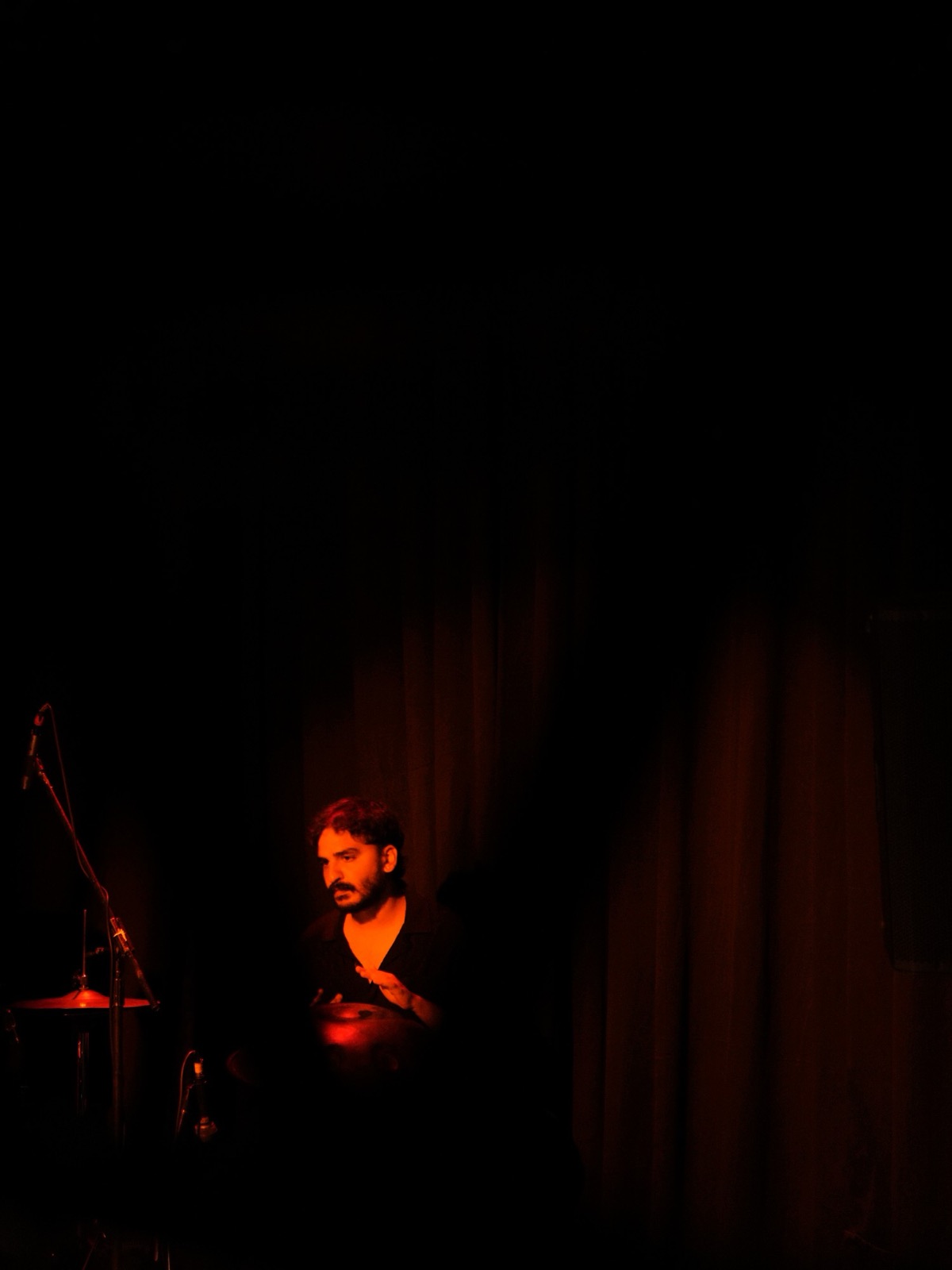 Ashim Bery alone in red light in front of a dark curtain, playing a handpan