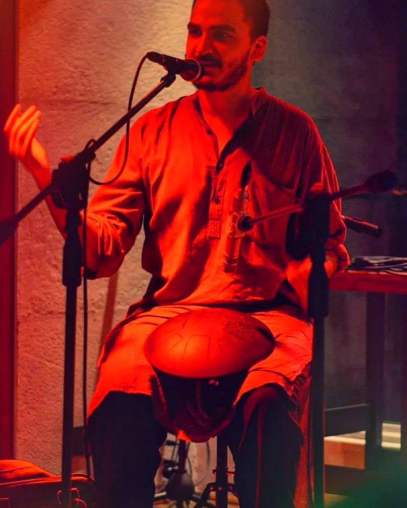 Ashim Bery in red light, speaking into a microphone with a tongue drum on his lap