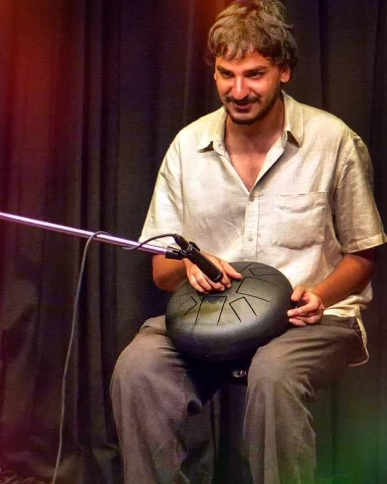 Ashim Bery smiling on stage with a steel tongue drum on his lap and a microphone in front