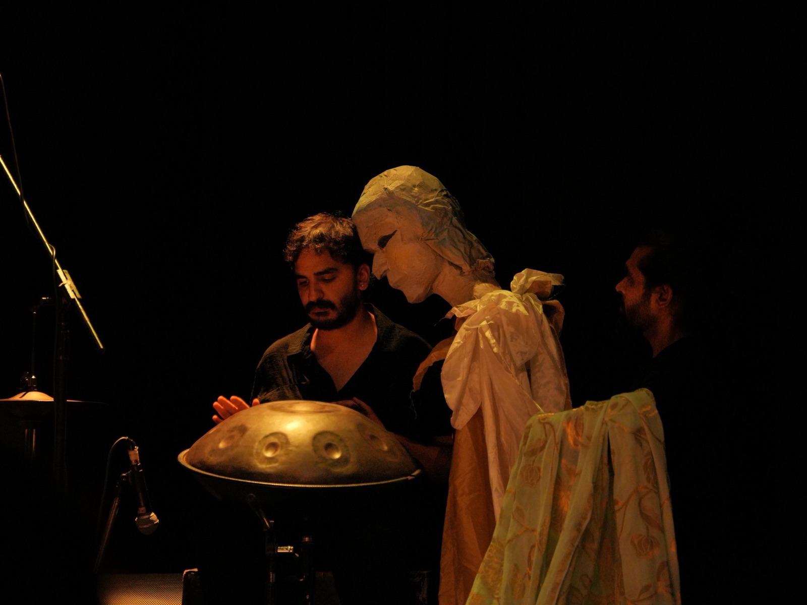 Ashim Bery playing a handpan on a dark stage in Spiral, a large masked puppet figure leaning in beside him